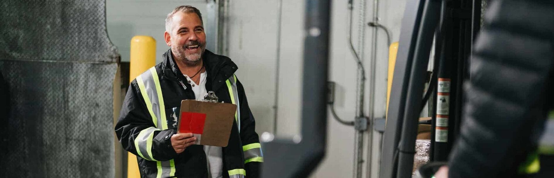 A warehouse worker smiling as he converses with a man operating a fork lift