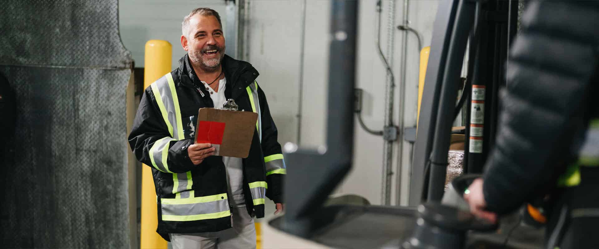 A warehouse worker smiling as he converses with a man operating a fork lift
